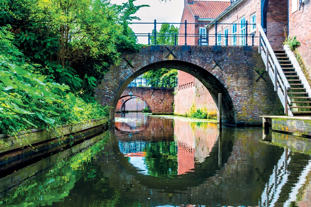A beautiful, serene water-level view looking down a narrow canal framed by old architecture and greenery. In the foreground, a prominent, semi-circular weathered red brick arch bridge spans across the calm water, creating a near-perfect reflection on the glassy canal surface. To the left, a wooden retaining wall holds back a lush bank covered in dense green leafy bushes and trees. To the right, a multi-story historic brick building meets the water's edge, featuring a wooden staircase leading up its side. In the background, a succession of two more brick arch bridges can be seen receding down the waterway.