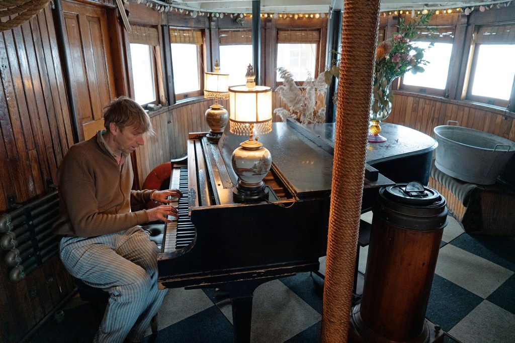 A man playing a black grand piano inside the wooden cabin saloon of a historic vessel, featuring vintage table lamps resting on the piano top, paneled walls, a checkered floor, and a rope-wrapped support pillar.