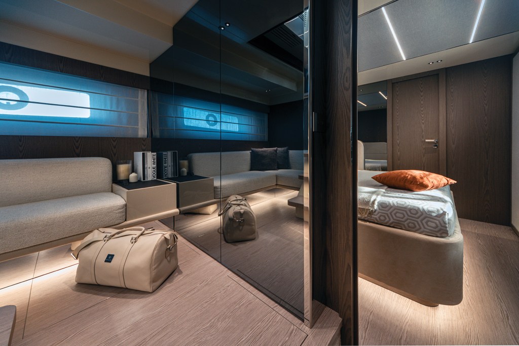 An interior shot of the ultra-modern, luxurious lower-deck master stateroom on an Invictus ST550 yacht. To the left, a cozy light grey sofa rests against dark wood paneling below a horizontal window with soft ambient lighting. A large, floor-to-ceiling smoked mirror wall divides the room, reflecting the seating area and a beige leather duffel bag on the light oak floor. To the right, a plush double bed with a geometric patterned coverlet and an orange accent pillow sits under integrated LED ceiling strips, positioned next to a matching wood-grain cabin door.