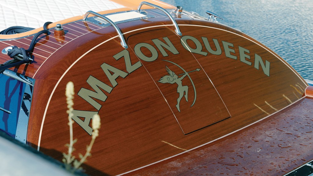 A close-up view of the rounded, tumblehome stern of the luxury motor yacht "Amazon Queen." The curved transom is crafted from highly polished, varnished mahogany wood and features the boat's name in bold, gold-colored capital letters arched over a custom graphic of an winged Amazon archer drawing a bow. The wooden swim platform below shows fresh water droplets, and stainless steel grab rails are visible at the top edge.