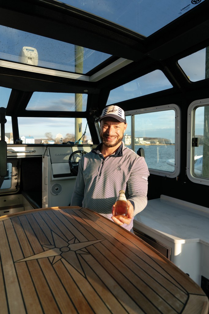 A medium shot from inside the bright, window-lined cabin of a Life Proof boat, featuring a smiling, bearded man holding out a small glass beer bottle. He wears a blue and white baseball cap and a grey striped long-sleeve shirt. In the foreground, a polished wooden table displays a large inlaid compass rose design. Sunlight streams through the cabin's large glass windows and overhead skylight panels, with calm open water visible in the background.