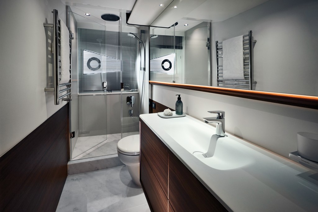 Luxurious modern ensuite head on a Princess V65 yacht featuring a white solid-surface vanity with an integrated sink, rich wood paneling, a marble-tiled floor, a toilet, and a glass-enclosed walk-in shower cubicle.