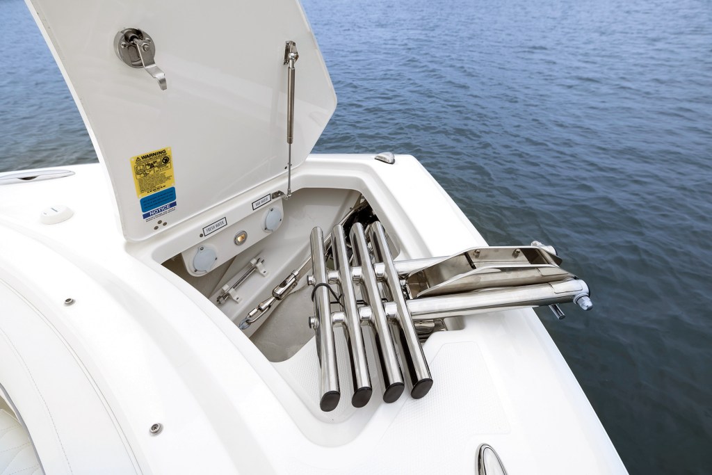 A close-up shot of the bow anchor locker on a Pursuit DC 286 boat, with the fiberglass hatch propped open by a stainless steel gas strut. Inside the recessed locker compartment, a telescoping stainless steel bow ladder is neatly folded and stowed, extending over the bow nose for beach boarding. The locker interior also reveals washdown connection inlets labeled "FRESH WATER" and "RAW WATER," set against a background of calm blue water.