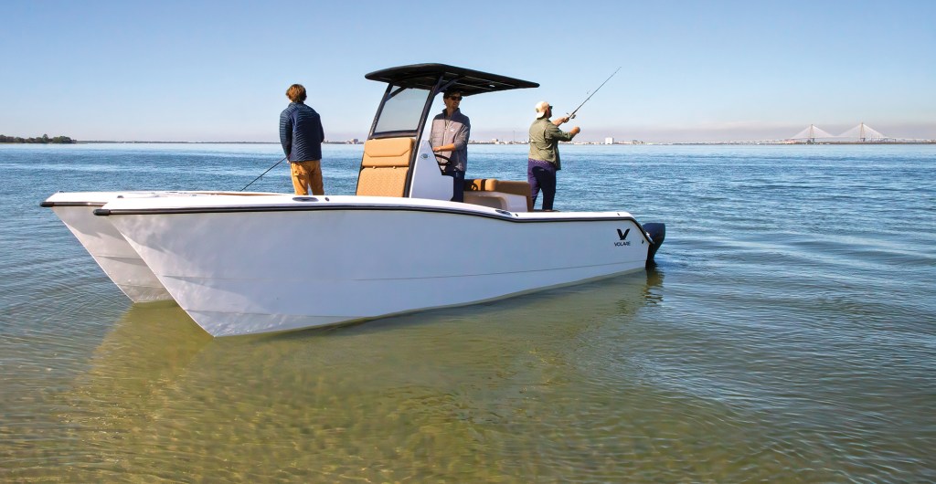 A wide, low-angle shot of a white Volare center-console power catamaran anchored in shallow, clear water near a sandy shoal. Three men are on board using the boat for fishing; one stands near the bow, another is at the helm beneath a black T-top canopy, and the third stands near the stern casting a fishing rod. The calm, blue water stretches out to a distant horizon where a large cable-stayed bridge is visible under a clear, bright sky.