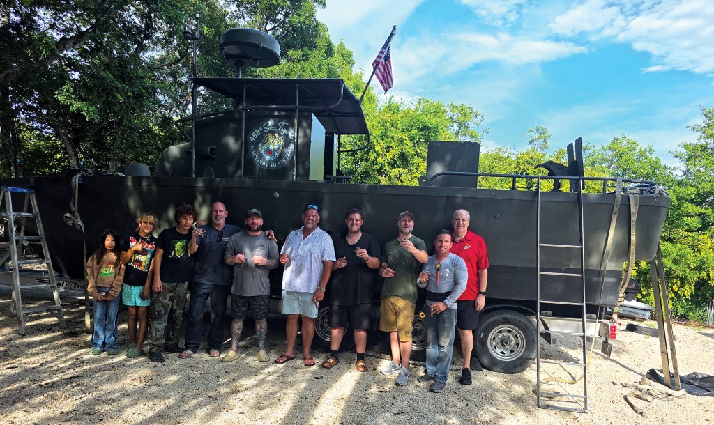 A wide, outdoor shot of a group of ten people posing for a group photo in front of a dark olive drab green military patrol boat on a trailer. The individuals, including children and adult men, stand shoulder-to-shoulder on a gravel surface under a bright, partly cloudy sky surrounded by green trees. The vessel features an American flag on the stern and a circular logo on the side that reads "458th Sea Tigers, Normandy to Vietnam." A silver ladder rests against the side of the boat, and an aluminum A-frame ladder stands nearby on the left.