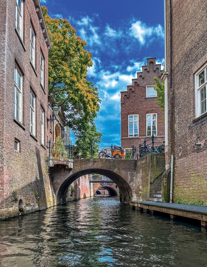 A vertical, low-angle shot from a rippling canal waterway framed tightly by tall, historic brick buildings on both sides. A weathered brick arch bridge connects the streets above, with a parked orange cargo bicycle visible on its span. To the right, a classic Dutch stepped-gable building rises against the sky, while a low wooden dock floats along the base of the brick wall below. Beyond the main bridge, a succession of smaller brick arches recedes down the canal route under a striking bright blue sky filled with white, wispy clouds.