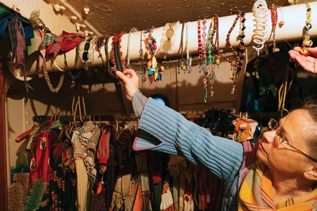 A woman with glasses browsing through a collection of colorful handmade necklaces and headbands hanging from an overhead pipe inside a boat's closet filled with vintage clothing.
