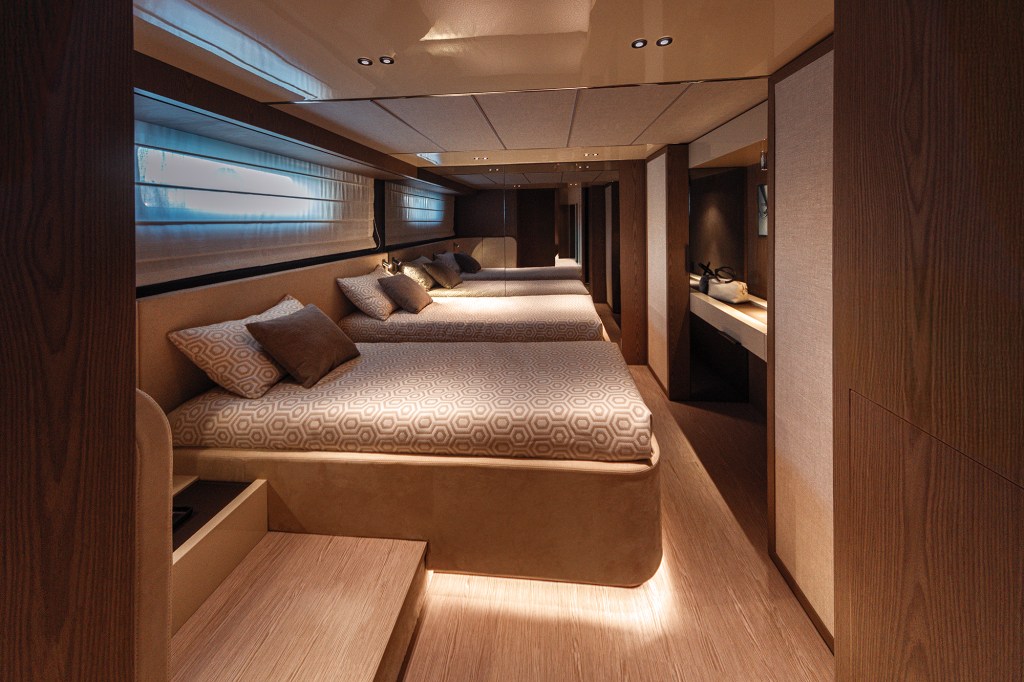 An interior shot of a spacious lower-deck guest stateroom on an Invictus ST550 yacht, featuring a modern warm-toned design. Two twin beds with geometric-patterned coverlets and brown pillows sit side-by-side on a light oak floor, enhanced by soft under-bed LED lighting. A large mirror wall behind the far bed reflects the room, adding to the sense of space. The left wall features a horizontal window with a soft white shade, while a floating vanity desk with a backlit mirror is visible on the right next to fine wood-grain paneling.