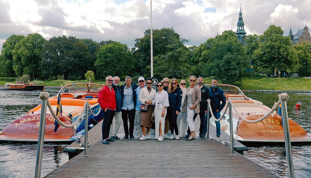 A wide, eye-level shot of a large group of people posing together on a wooden dock between two classic motor yachts. To the left is the yacht "Amazon Queen" with its distinctive orange upholstery, and to the right is a matching wood-trimmed yacht named "Zens." Lush green trees line the background waterway under a partly cloudy sky, with a historic European-style spire visible behind the treeline on the right.