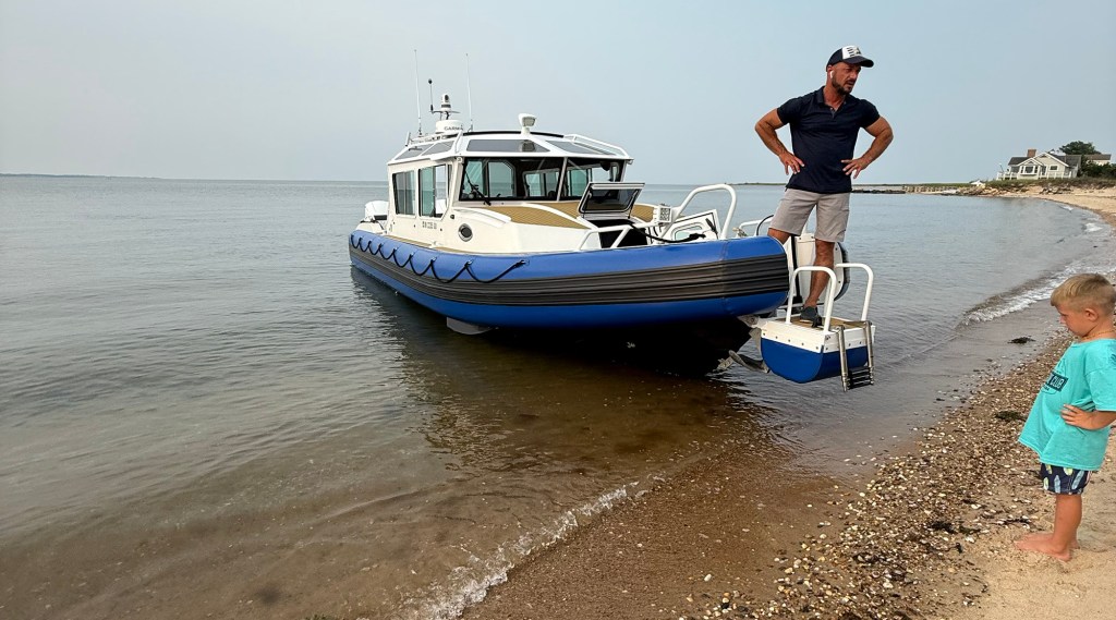 A wide, eye-level shot of a white Life Proof rigid inflatable boat (RIB) with a blue collar hull nose-in onto a calm sandy beach. A man stands at the bow door with hands on his hips, looking down toward a young boy in a turquoise t-shirt standing on the pebbled shoreline. The boat features an enclosed cabin cruiser design, an open bow hatch, and a custom drop-down bow door with a white metal boarding ladder extension. A single coastal house sits on a grassy bluff in the distant right background under an overcast sky.