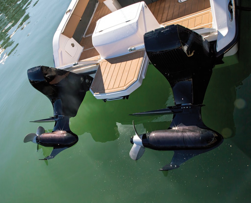 A close-up, high-angle shot looking down at the stern transom and twin black outboard motor lower units on a white Volare catamaran. The glossy black engine legs and gearcases extend down into clear, light-green water, showing their stainless steel propellers reflecting the bright sunlight. Between the two engines, a light brown faux-teak swim platform extension sits below a white storage compartment box on the cockpit deck.