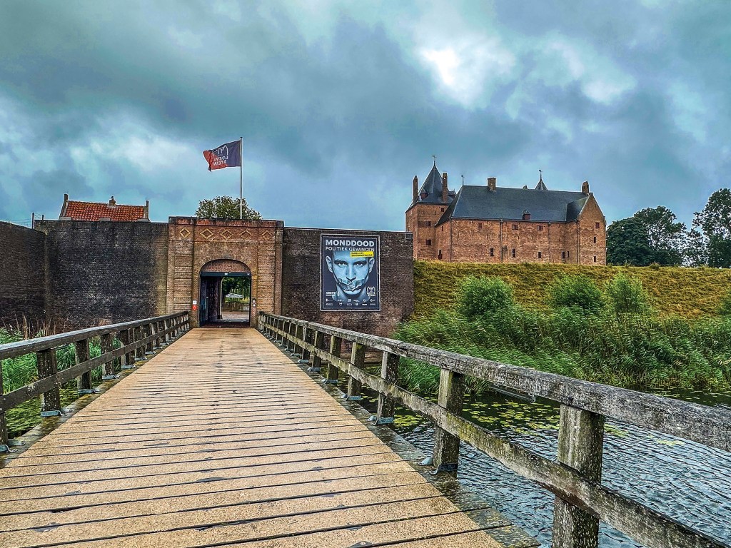 A wide, straight-on view of a wooden plank bridge with rustic timber railings leading across a water-filled moat toward a historic stone fortress wall. An arched gateway entry penetrates the fortified wall on the left, topped with a flying flag. To the right of the gate, a large black informational poster featuring a face is mounted on the wall. Rising on a grassy hill behind the fortifications stands Loevestein Castle, a multi-story medieval brick fortress with pointed roofs, set under a heavy, dramatic overcast sky.
