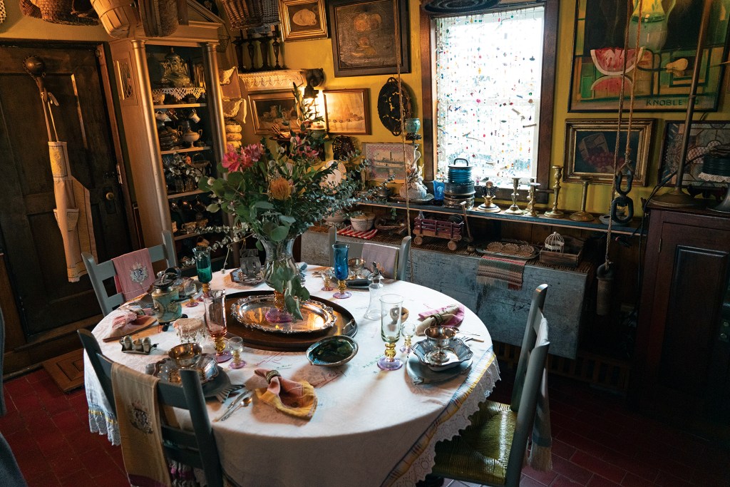An eclectic dining room setup with a round table beautifully set with vintage glassware, silver platters, and a large floral centerpiece, surrounded by walls decorated with paintings, trinkets, and a beaded window curtain.