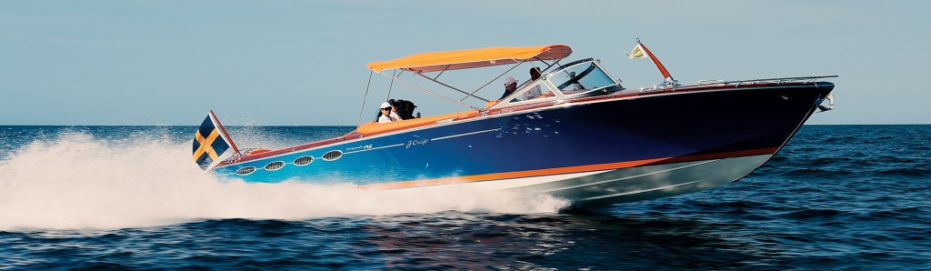 A wide panoramic, profile shot of the luxury motor yacht "Amazon Queen" cruising at high speed across a deep blue ocean. The sleek Prussian blue hull cuts through the water, throwing up a dramatic white wake and sea spray from its stern, where a Swedish flag flies proudly. An orange bimini top is deployed over the open cockpit to provide shade for the passengers and driver as the boat moves swiftly under a clear sky.