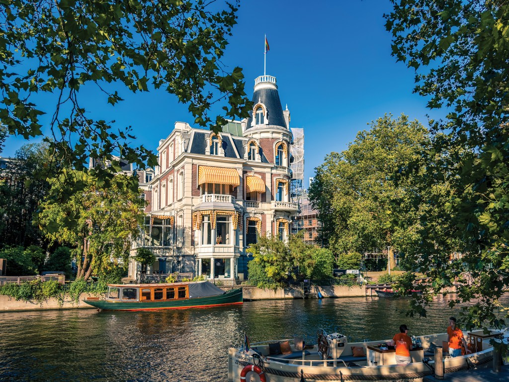 A sunny, wide-angle shot looking across a glittering canal at an elegant, multi-story historic mansion. The building features ornate white trim, a dark mansard roof, yellow-and-white striped window awnings, and a distinctive rounded corner tower topped with a flagpole. A traditional wooden canal boat with a green hull is moored along the stone canal wall in front of the house. In the foreground, the bow of another boat with passengers in orange shirts is visible, while lush green leafy trees frame the entire scene against a clear, deep blue sky.