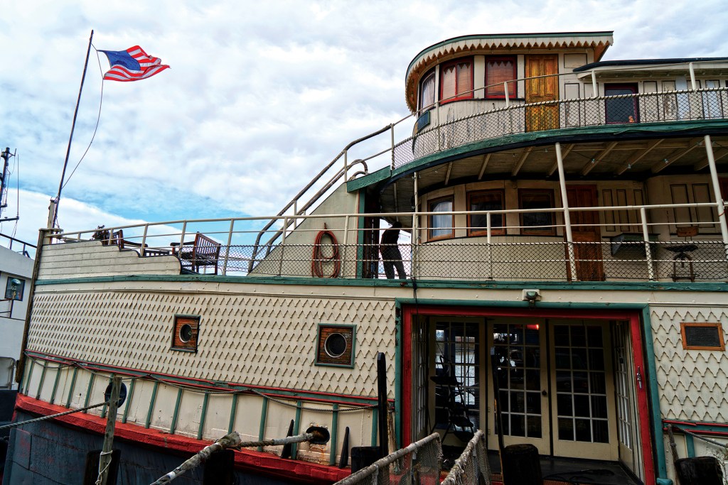 Side exterior profile of the historic wooden ferryboat Yankee docked at a pier, showcasing its multi-deck architecture, patterned hull shingles, glass entryway doors, and an American flag waving from the bow under a cloudy sky.