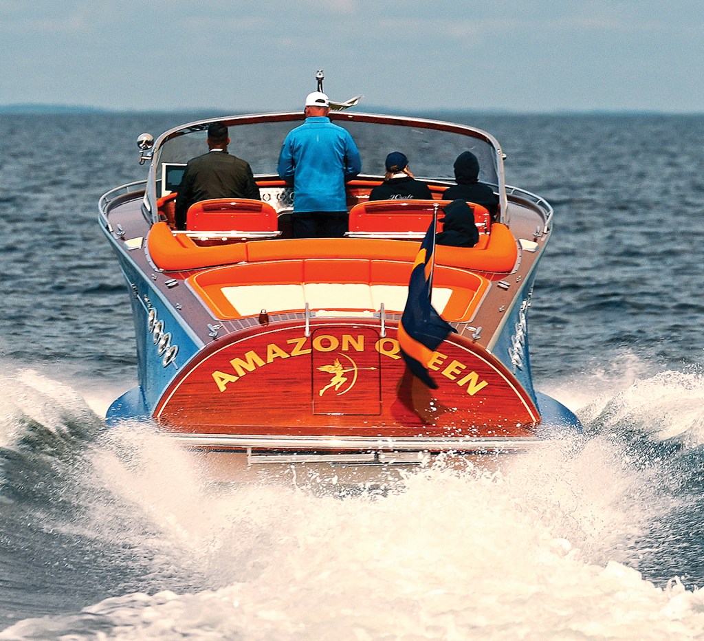 A rear, eye-level shot capturing the luxury yacht "Amazon Queen" underway on open water from directly behind the stern. The vibrant orange cockpit seating is occupied by four passengers, including the captain standing centered at the wheel in a blue jacket. Heavy white water spray and a churning wake erupt from beneath the polished mahogany transom, which displays the gilded boat name, custom archer logo, and an attached flag fluttering in the wind.