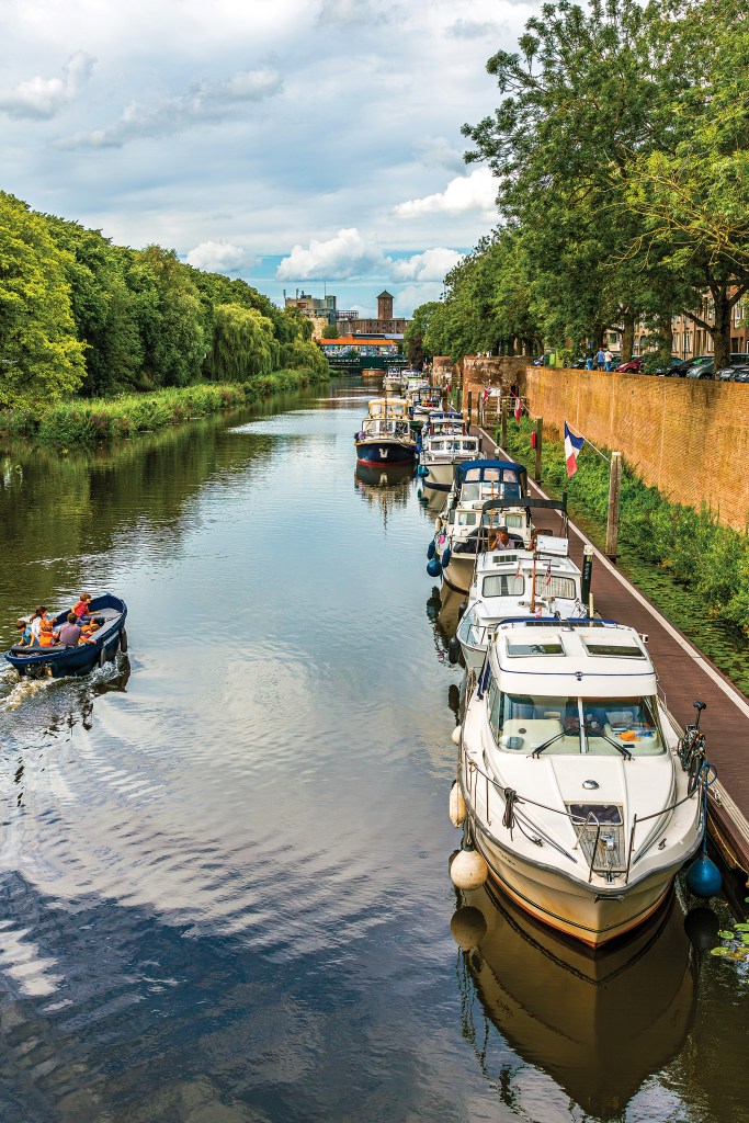 A high-angle, vertical perspective looking down a long, calm canal lined with lush greenery and boats. Along the right bank, a wooden floating dock is packed with a neat row of moored motor yachts and cabin cruisers. This side of the canal is bordered by a tall, linear red brick retaining wall topped with trees, where a small French flag flies from a wooden post. On the left side of the canal, a small blue open motorboat carrying several passengers cruises down the waterway, leaving a gentle wake. The water creates soft, glassy reflections of the boats and the partly cloudy blue sky.