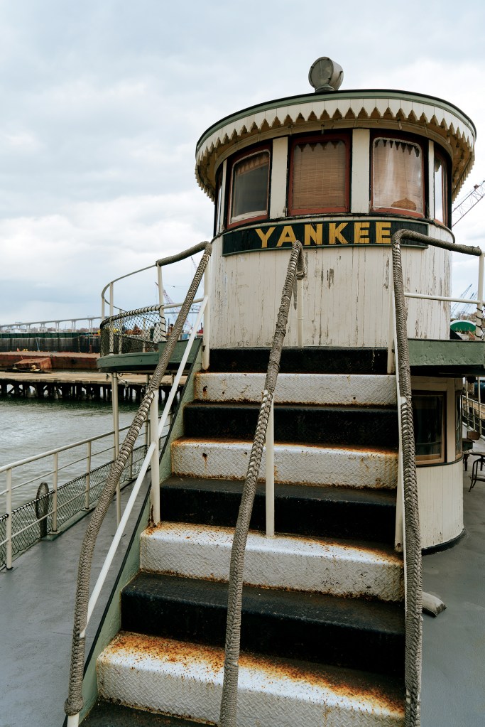 A set of weathered metal stairs with rope handrails leading up to the rounded pilothouse of the historic ferryboat Yankee, showing signs of rust on the steps and an industrial harbor background.