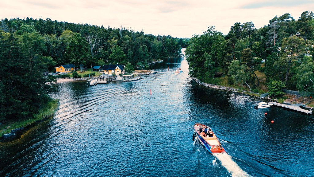 An aerial, high-angle drone shot capturing the luxury yacht "Amazon Queen" as it navigates down a narrow, scenic inland waterway channel. The deep blue water is flanked on both sides by dense green pine and deciduous forests. Yellow and dark-trimmed waterfront cottages with private docks sit on the left shoreline, while another small motorboat can be seen cruising further ahead in the distance.