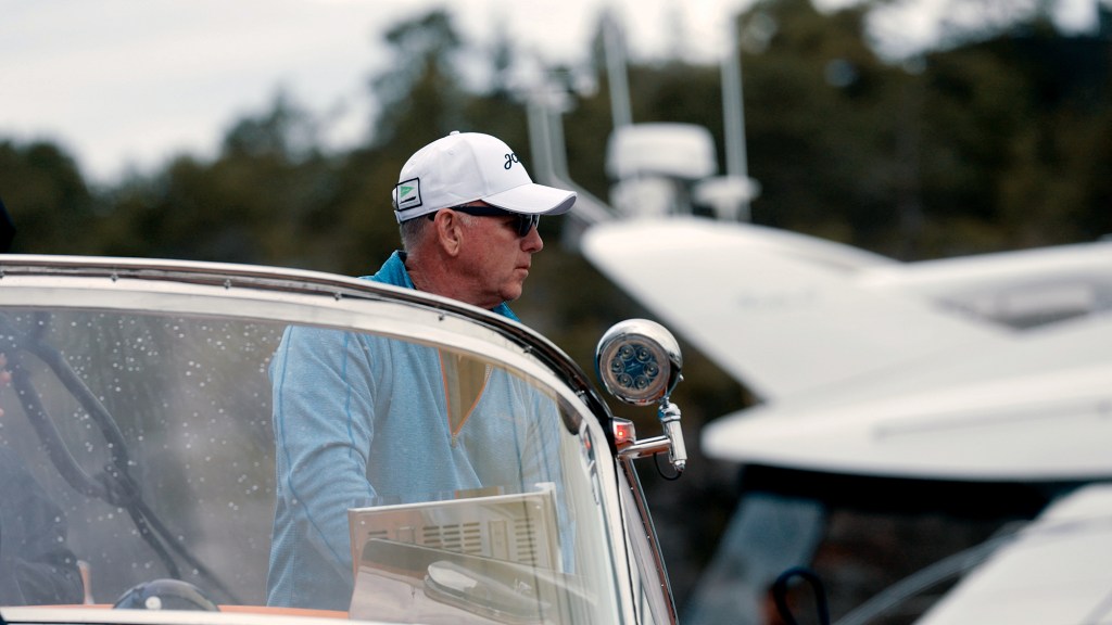 A medium close-up, profile shot of a man wearing a white baseball cap and sunglasses at the helm of a boat. He is dressed in a light-blue, quarter-zip pullover shirt and is looking intently forward behind the boat's curved glass windshield, which has water droplets on its surface. A round, chrome-plated spotlight is mounted near the edge of the helm structure, with a blurred white boat superstructure and green trees visible in the soft-focus background.