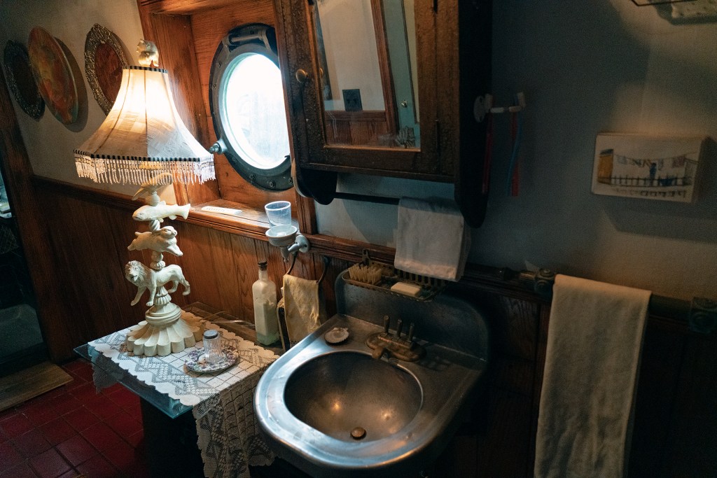 A vintage boat head interior featuring a classic stainless steel basin sink, a wooden medicine cabinet mirror, a round nautical porthole window, and an ornate table lamp with animal carvings resting on a lace doily next to the sink.