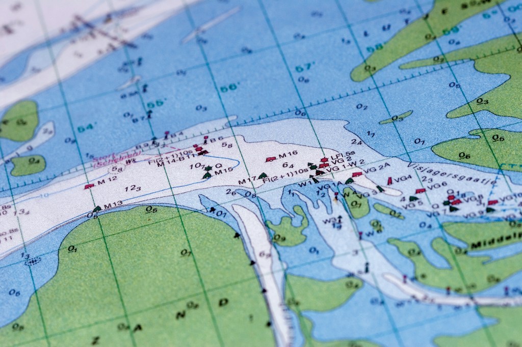A detailed overhead view of a printed marine navigation chart showing coastal waterways, shallow sandbanks, and channels. The map uses blue for water depth, green for landmasses, and includes various nautical symbols, numbers for water depth coordinates, and textual labels.