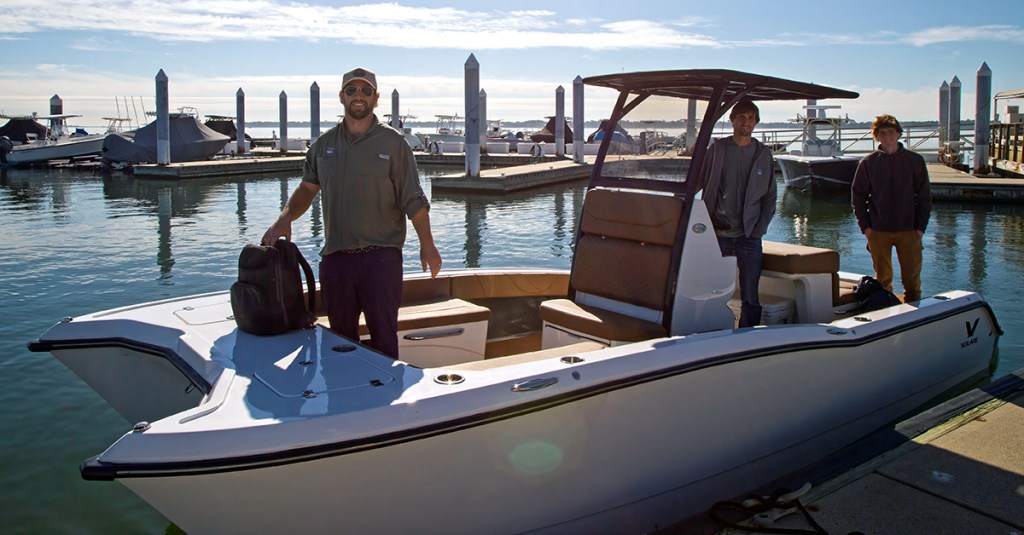 A wide shot of three men on board a white Volare center-console power catamaran docked at a sunny marina. In the foreground, a man wearing a baseball cap stands on the open bow deck holding a black backpack. A second man stands at the helm station beneath a black T-top canopy, while a third man stands near the stern deck on the right. The boat features tan upholstered seating and a prominent black "V" logo on its aft hull, set against a background of calm water, concrete docks, and vertical pilings under a bright sky.