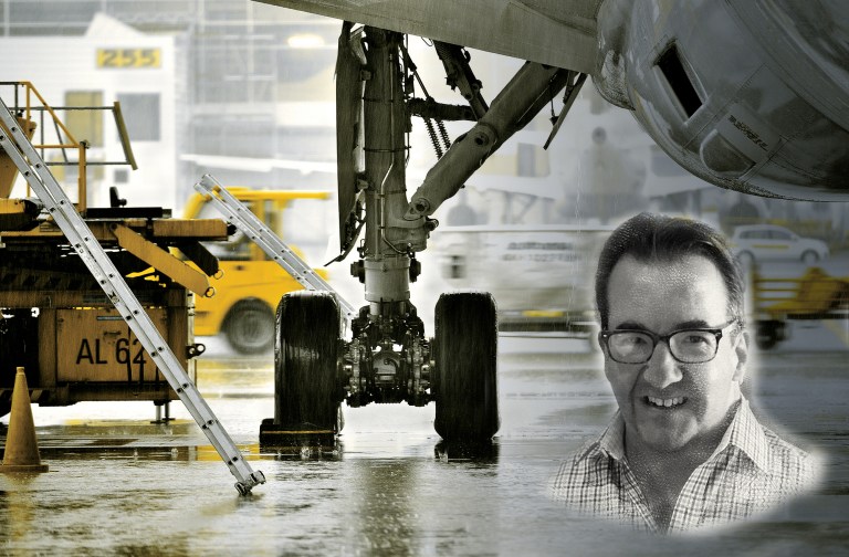 A stylized, low-angle composite image set on a wet, rainy airport tarmac, utilizing a desaturated color palette with selective yellow highlights. In the center, the heavy landing gear assembly and dual tires of a large commercial aircraft rest on the reflective ground. To the left, yellow ground support equipment, a ladder, and a traffic cone are visible. Superimposed in the lower-right corner is a black-and-white portrait of a smiling middle-aged man wearing glasses and a plaid button-down shirt.