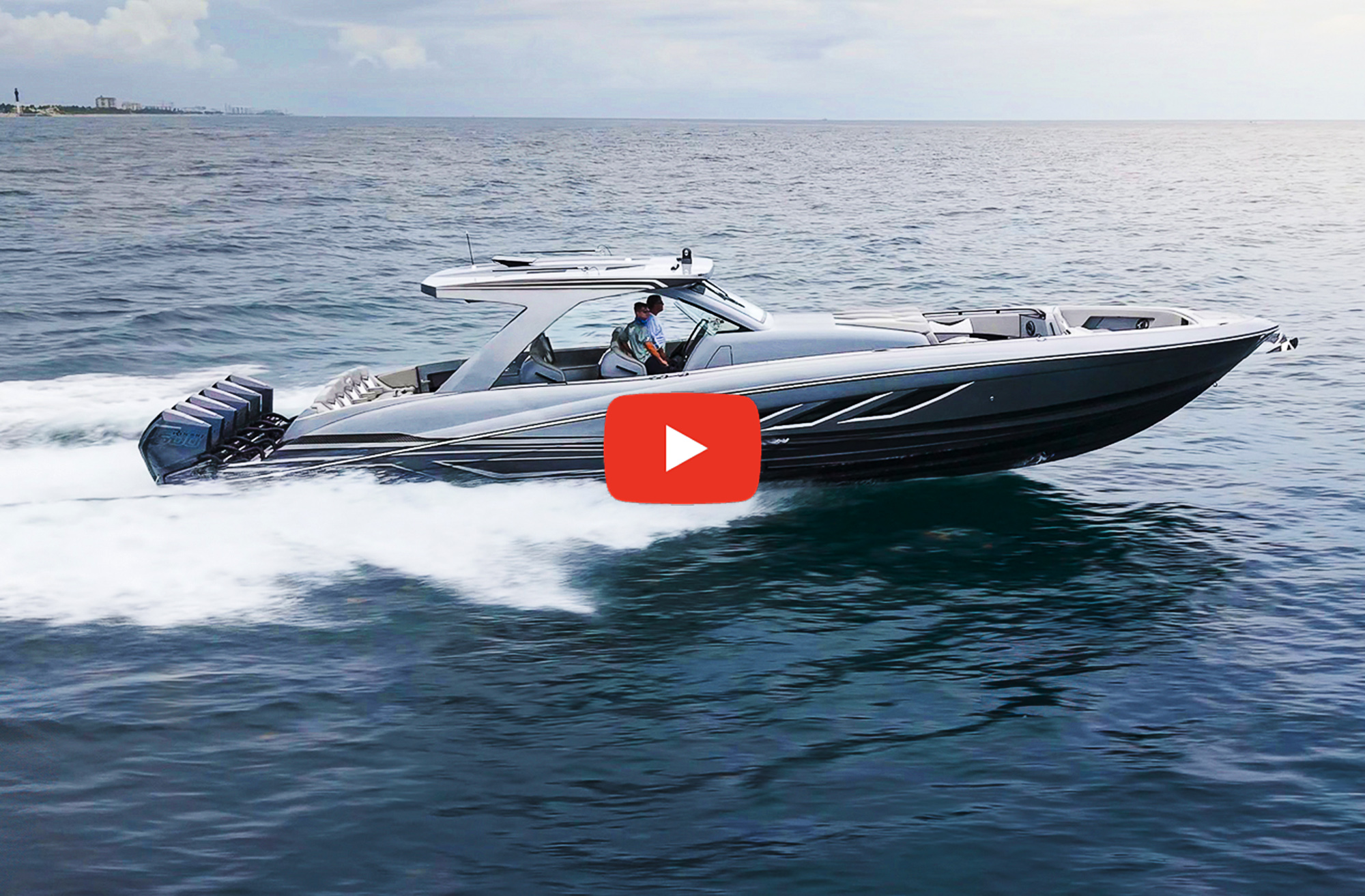 A high-angle view of a grey and black Deep Impact 499 Sport luxury center-console boat cruising fast across dark blue coastal waters, leaving a white wake. Two people stand at the helm beneath a hardtop canopy, and a line of six outboard motors is mounted on the transom. A prominent red YouTube play button icon is overlaid in the center of the image, framing the boat against a calm sea and a hazy horizon under an overcast sky.