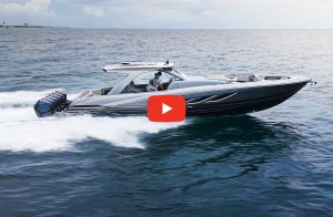 feat-Deep-Imact-499-Sport A high-angle view of a grey and black Deep Impact 499 Sport luxury center-console boat cruising fast across dark blue coastal waters, leaving a white wake. Two people stand at the helm beneath a hardtop canopy, and a line of six outboard motors is mounted on the transom. A prominent red YouTube play button icon is overlaid in the center of the image, framing the boat against a calm sea and a hazy horizon under an overcast sky.