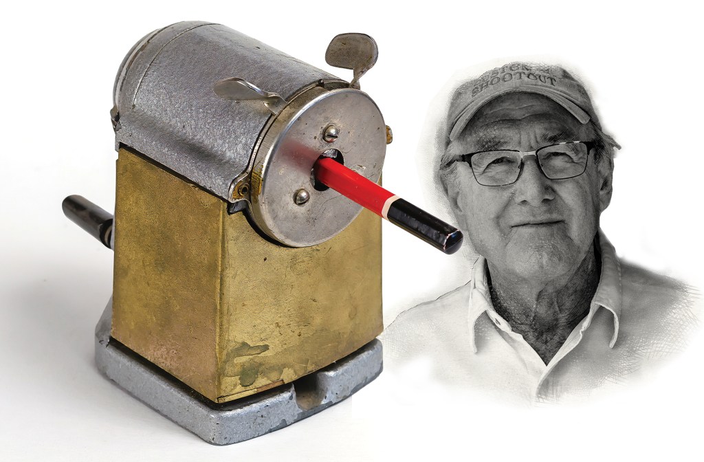 A studio composite image on a plain white background. On the left is an antique mechanical pencil sharpener featuring a square brass-colored body, a distressed silver metal curved top with a hand crank handle, and two small wing tabs on top. A red pencil with a black end is inserted into its circular front guide. On the right, a black-and-white portrait vignette shows an elderly man smiling gently, wearing a baseball cap and glasses.