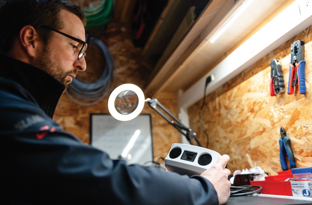 A close-up profile shot of a male technician with glasses working on electronic hardware at a well-lit workshop bench. He focuses intently while holding a compact white optical housing unit equipped with dual lenses and a central camera sensor. A bright, illuminated desktop ring magnifying lamp sits on an adjustable arm in the center. In the background, hand tools hang neatly on a wooden pegboard wall beneath a bright LED under-cabinet light bar, with wire spools coiled overhead.