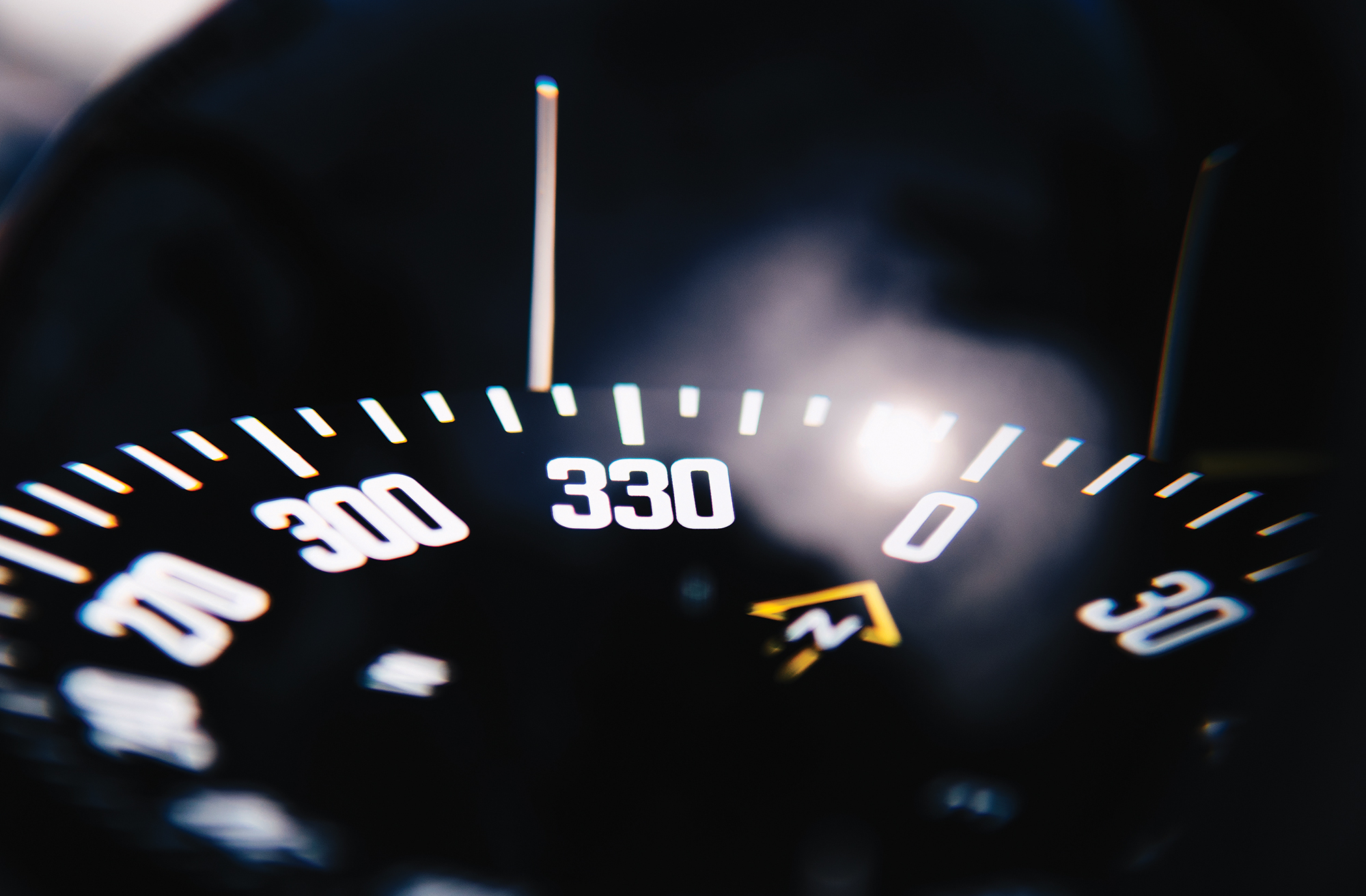 A close-up, selective-focus shot of a black marine compass dial illuminated at night. Bold white numbers indicate bearings for 300, 330, and 0 (North), with a yellow arrow symbol pointing toward the North marker beneath a bright reflection on the glass.