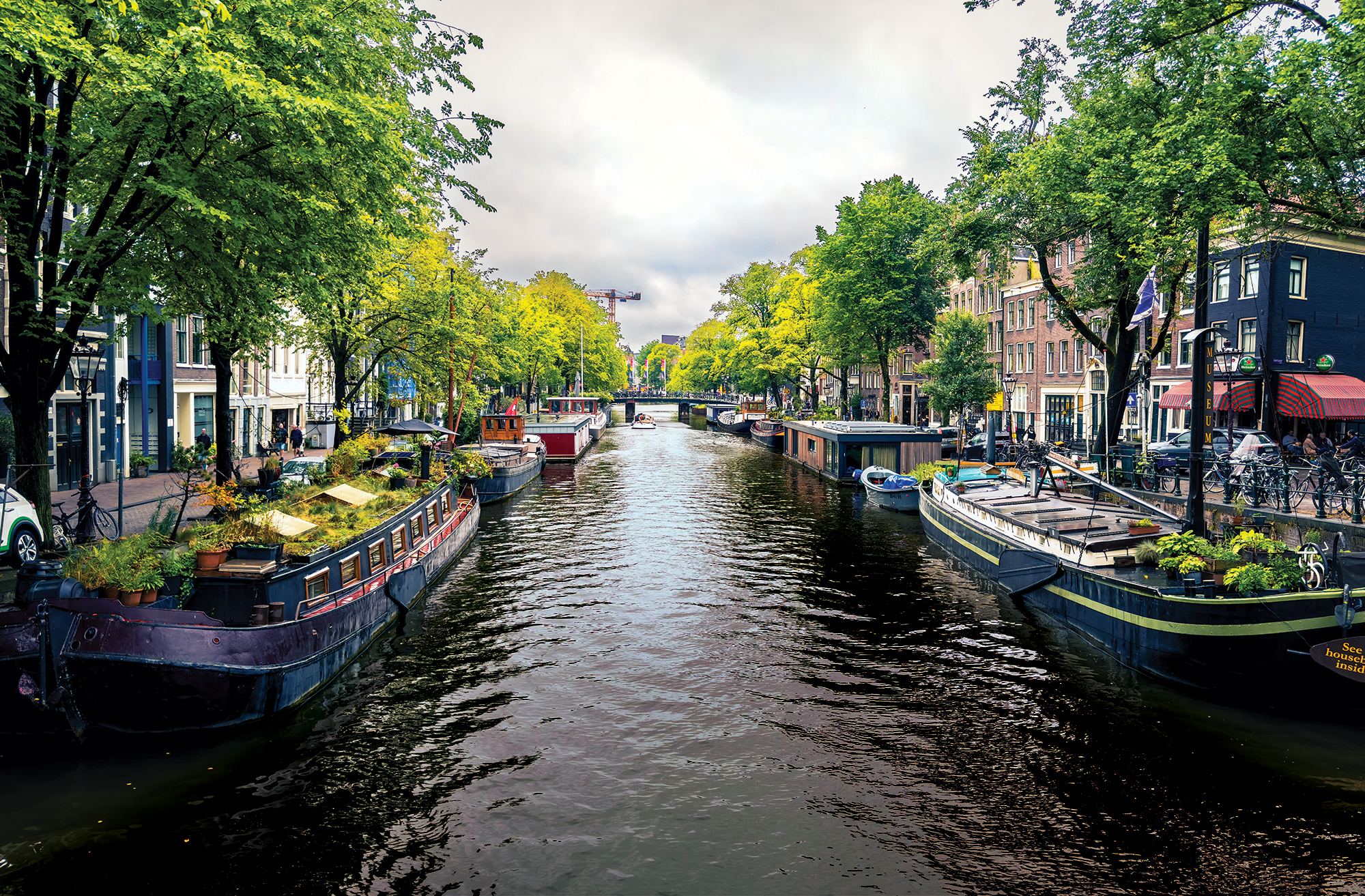 A scenic, eye-level perspective looking down a busy historic canal waterway in Amsterdam. Long, traditional houseboats line both sides of the canal banks, some featuring lush container gardens and potted plants on their roofs. The water reflects the cloudy sky above. Dense rows of vibrant green trees frame the canal, partially shading cobblestone streets, brick townhomes, parked bicycles, and a cafe terrace on the right. In the distance, a low arched bridge spans across the water under an overcast, dramatic sky.