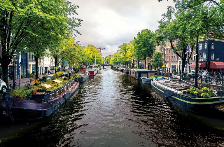 A scenic, eye-level perspective looking down a busy historic canal waterway in Amsterdam. Long, traditional houseboats line both sides of the canal banks, some featuring lush container gardens and potted plants on their roofs. The water reflects the cloudy sky above. Dense rows of vibrant green trees frame the canal, partially shading cobblestone streets, brick townhomes, parked bicycles, and a cafe terrace on the right. In the distance, a low arched bridge spans across the water under an overcast, dramatic sky.