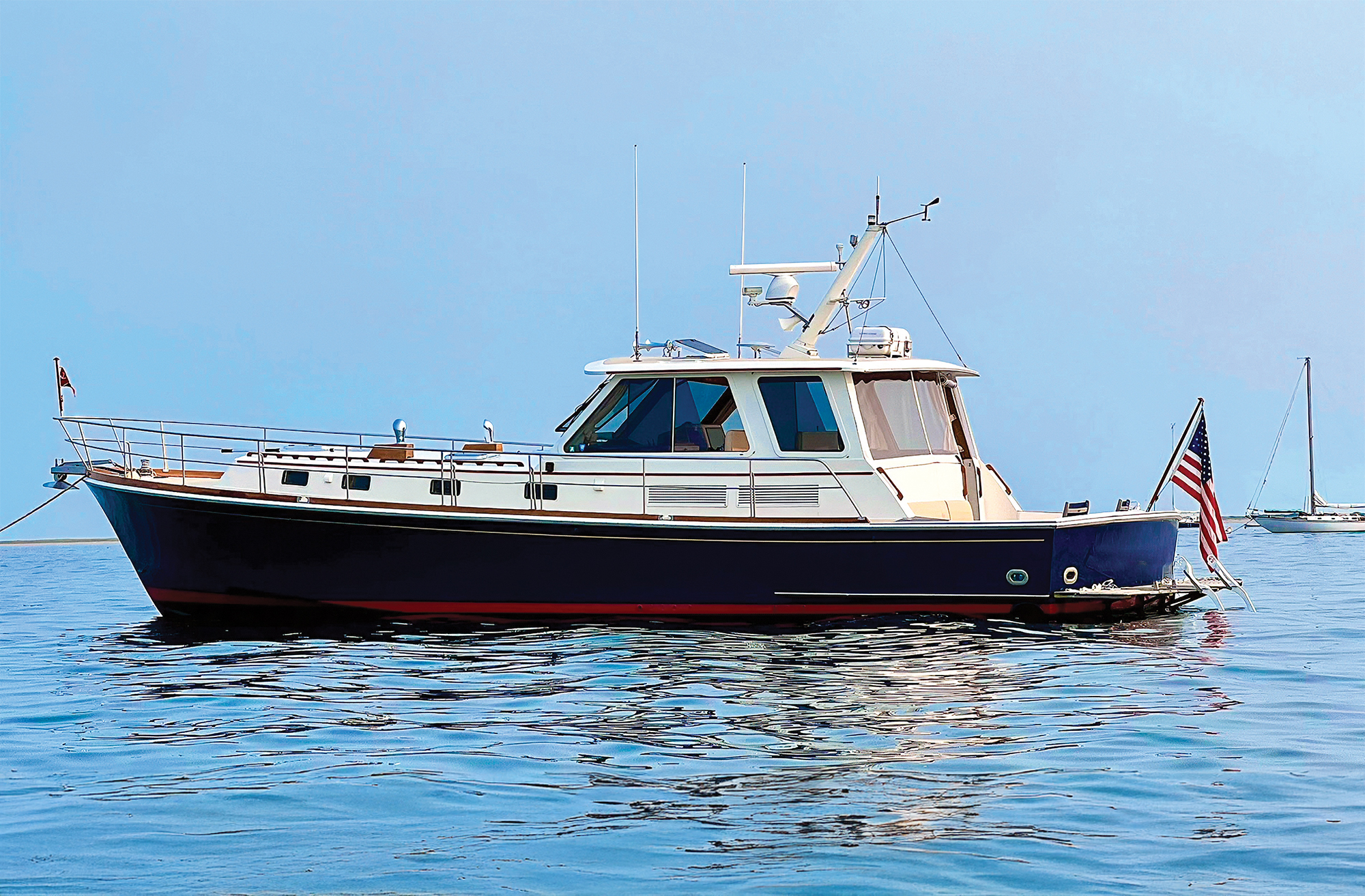 A profile shot of a classic dark blue and white Downeast-style yacht floating on calm, glass-like blue water under a clear sky. The vessel features an enclosed pilothouse with tinted glass windows, traditional teak wood accents along the deck line, a radar mast on the hardtop roof, and an American flag flying at the stern. A distant sailboat is subtly visible anchored on the horizon to the right.