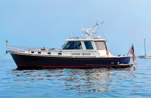 A profile shot of a classic dark blue and white Downeast-style yacht floating on calm, glass-like blue water under a clear sky. The vessel features an enclosed pilothouse with tinted glass windows, traditional teak wood accents along the deck line, a radar mast on the hardtop roof, and an American flag flying at the stern. A distant sailboat is subtly visible anchored on the horizon to the right.