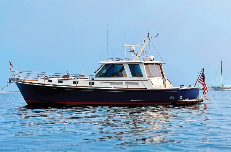 A profile shot of a classic dark blue and white Downeast-style yacht floating on calm, glass-like blue water under a clear sky. The vessel features an enclosed pilothouse with tinted glass windows, traditional teak wood accents along the deck line, a radar mast on the hardtop roof, and an American flag flying at the stern. A distant sailboat is subtly visible anchored on the horizon to the right.