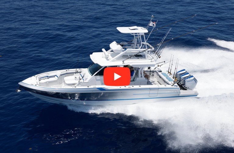 feat_Formula-387-CCF An aerial, high-angle action shot of a white and metallic blue Formula 387 Center Console Fish (CCF) yacht cruising fast across a deep blue ocean. The high-performance boat features a large t-top hardtop with an upper helm station, outriggers extended for fishing, and a row of fishing rods mounted in the cockpit. It is powered by triple white Mercury outboard motors, throwing a large white spray of water and wake behind it. A large red YouTube-style play button icon is overlaid directly in the center of the image.
