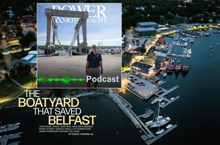 An aerial, high-angle twilight view of a bustling waterfront shipyard and marina with a prominent article title overlay reading "THE BOATYARD THAT SAVED BELFAST." In the top left corner, a rectangular inset displays a Power & Motoryacht podcast graphic showing a man standing in front of a massive "FRONT STREET SHIPYARD" mobile boat hoist, complete with a green audio waveform and the word "Podcast." The background shows the illuminated Safe Harbor Front Street Shipyard facility in Belfast, Maine, with docks, service buildings, and boats floating in the calm water.