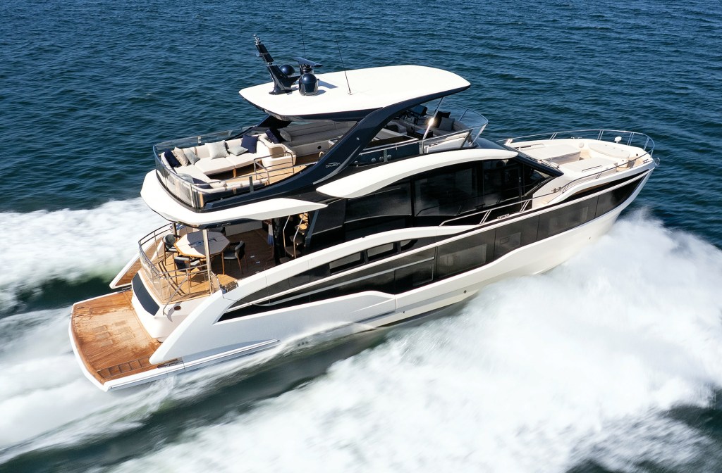 A high-angle, overhead aerial action shot of a sleek white and black Galeon 620 Fly luxury yacht cruising fast through dark blue ocean water. The three-deck flybridge vessel carves a sharp turn, generating a dramatic white spray and wake along its starboard side. The massive upper flybridge features extensive beige outdoor sofa seating under a white hardtop bimini roof with black support arches and radar equipment. Below, the teak aft deck cockpit shows a dining table setup, and large dark-tinted glass windows line the main salon deck.