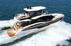 A high-angle, overhead aerial action shot of a sleek white and black Galeon 620 Fly luxury yacht cruising fast through dark blue ocean water. The three-deck flybridge vessel carves a sharp turn, generating a dramatic white spray and wake along its starboard side. The massive upper flybridge features extensive beige outdoor sofa seating under a white hardtop bimini roof with black support arches and radar equipment. Below, the teak aft deck cockpit shows a dining table setup, and large dark-tinted glass windows line the main salon deck.