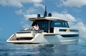 A low-angle, three-quarter view from the water looking up at a white Invictus ST55 luxury motor yacht idling on calm blue seas. A couple sits relaxing on the spacious aft sunpad deck outside the enclosed main cabin, which features dark tinted windows and black radar domes on the roof. The yacht's modern design includes a fold-down bulwark terrace extended on the starboard side just above the waterline, set against a bright blue sky filled with fluffy white clouds.