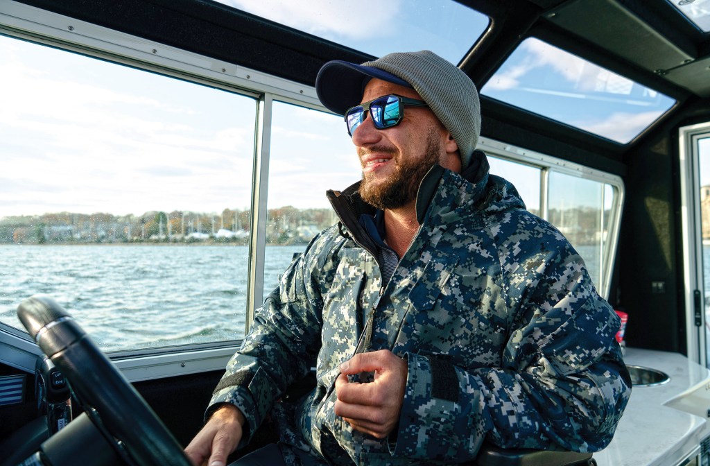 A medium shot from inside an enclosed boat helm station featuring a smiling, bearded man sitting at the steering wheel. He is looking off to the right, wearing blue-mirrored sunglasses, a blue baseball cap underneath a grey beanie, and a hooded blue digital-camouflage jacket. Large glass windows and an overhead skylight panel surround the cabin, offering a view out over choppy water toward a distant, tree-lined shoreline under a bright sky.