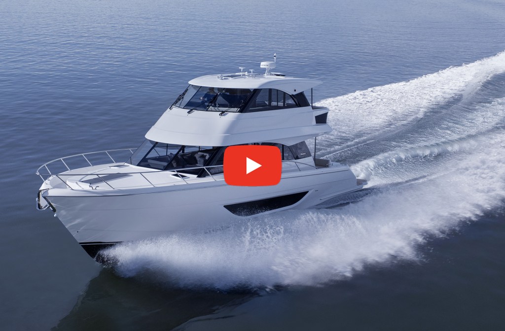 A high-angle, three-quarter view of a sleek, white Maritimo M50 luxury flybridge motor yacht cruising swiftly across a calm blue sea. The boat cuts sharply through the water, throwing up a bright white spray along its hull and leaving a long, foaming wake behind it. A large, red YouTube-style play button icon is centered over the middle of the yacht, framing the vessel against the wide open ocean.