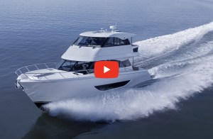 A high-angle, three-quarter view of a sleek, white Maritimo M50 luxury flybridge motor yacht cruising swiftly across a calm blue sea. The boat cuts sharply through the water, throwing up a bright white spray along its hull and leaving a long, foaming wake behind it. A large, red YouTube-style play button icon is centered over the middle of the yacht, framing the vessel against the wide open ocean.