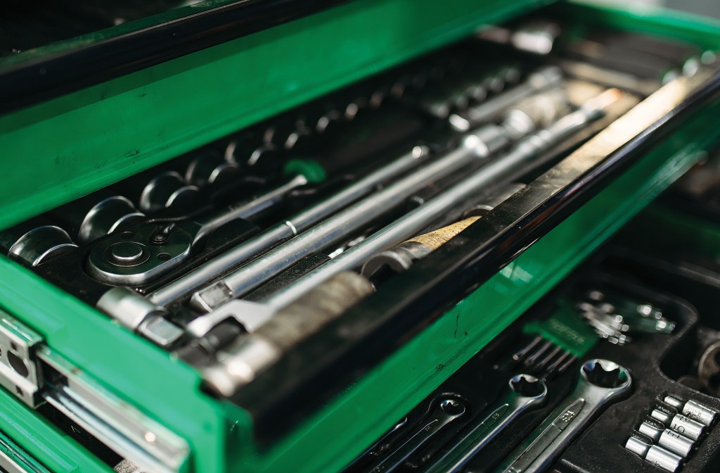 A close-up, high-angle shot looking into the open drawers of a bright green mechanic's toolbox filled with tools. The shallow top drawer is in sharp focus, neatly organizing a socket wrench ratchet, long chrome extension bars, and a row of round sockets. The drawer below it is slightly out of focus, revealing open-end wrenches, hex keys, and additional smaller sockets nestled in black foam tray inserts.
