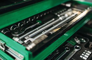 A close-up, high-angle shot looking into the open drawers of a bright green mechanic's toolbox filled with tools. The shallow top drawer is in sharp focus, neatly organizing a socket wrench ratchet, long chrome extension bars, and a row of round sockets. The drawer below it is slightly out of focus, revealing open-end wrenches, hex keys, and additional smaller sockets nestled in black foam tray inserts.