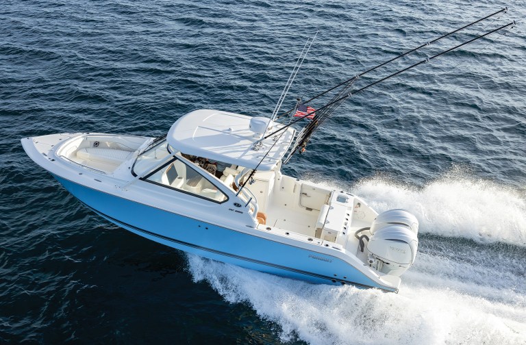 feat_Pursuit DC 286 An aerial, high-angle shot of a light-blue and white Pursuit DC 286 dual-console sports boat cruising fast through dark blue choppy waters. The boat features an open bow seating area, a white hardtop with a radar dome, and twin white Yamaha outboard motors producing a heavy white wake. Long black outrigger poles extend backward from the hardtop canopy, framing the fishing-equipped boat as it cuts through the open ocean.